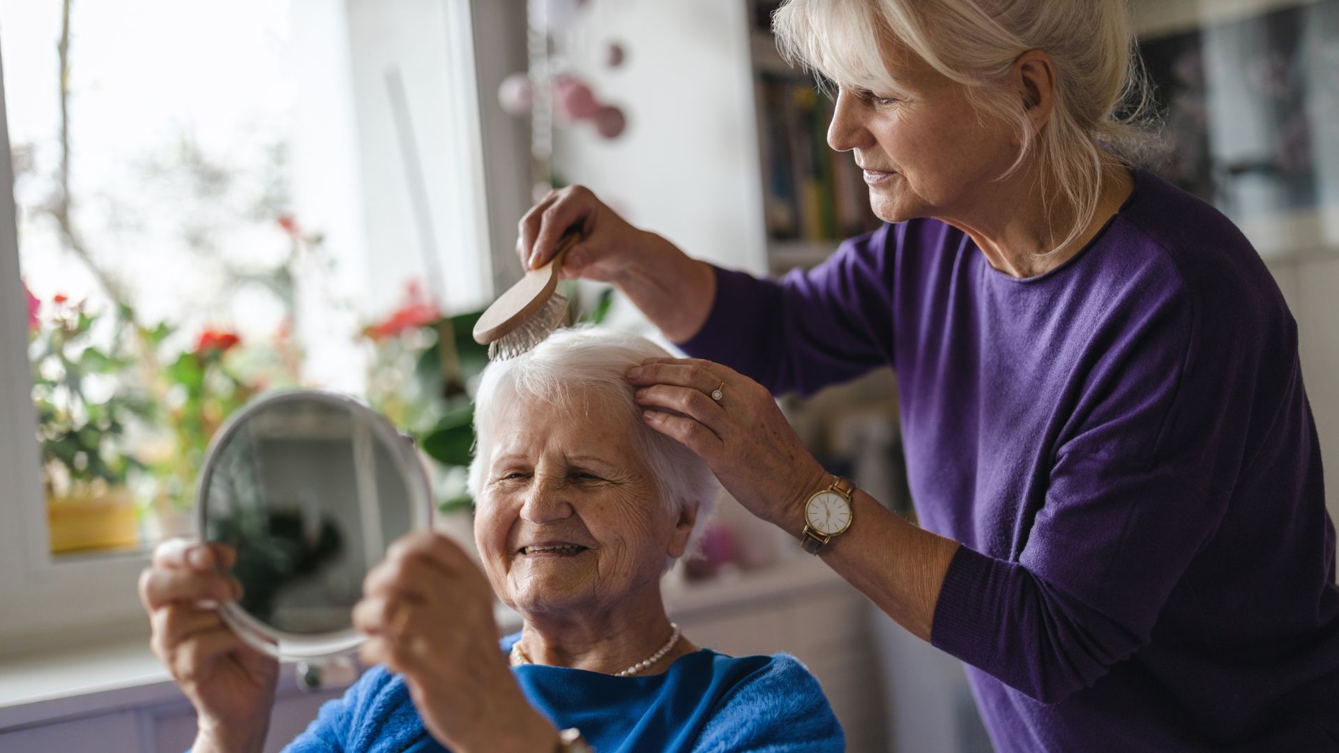 An older woman gently brushes the hair of another smiling elderly woman who is holding a mirror, capturing a tender caregiving moment often experienced in families affected by Alzheimer’s Disease.