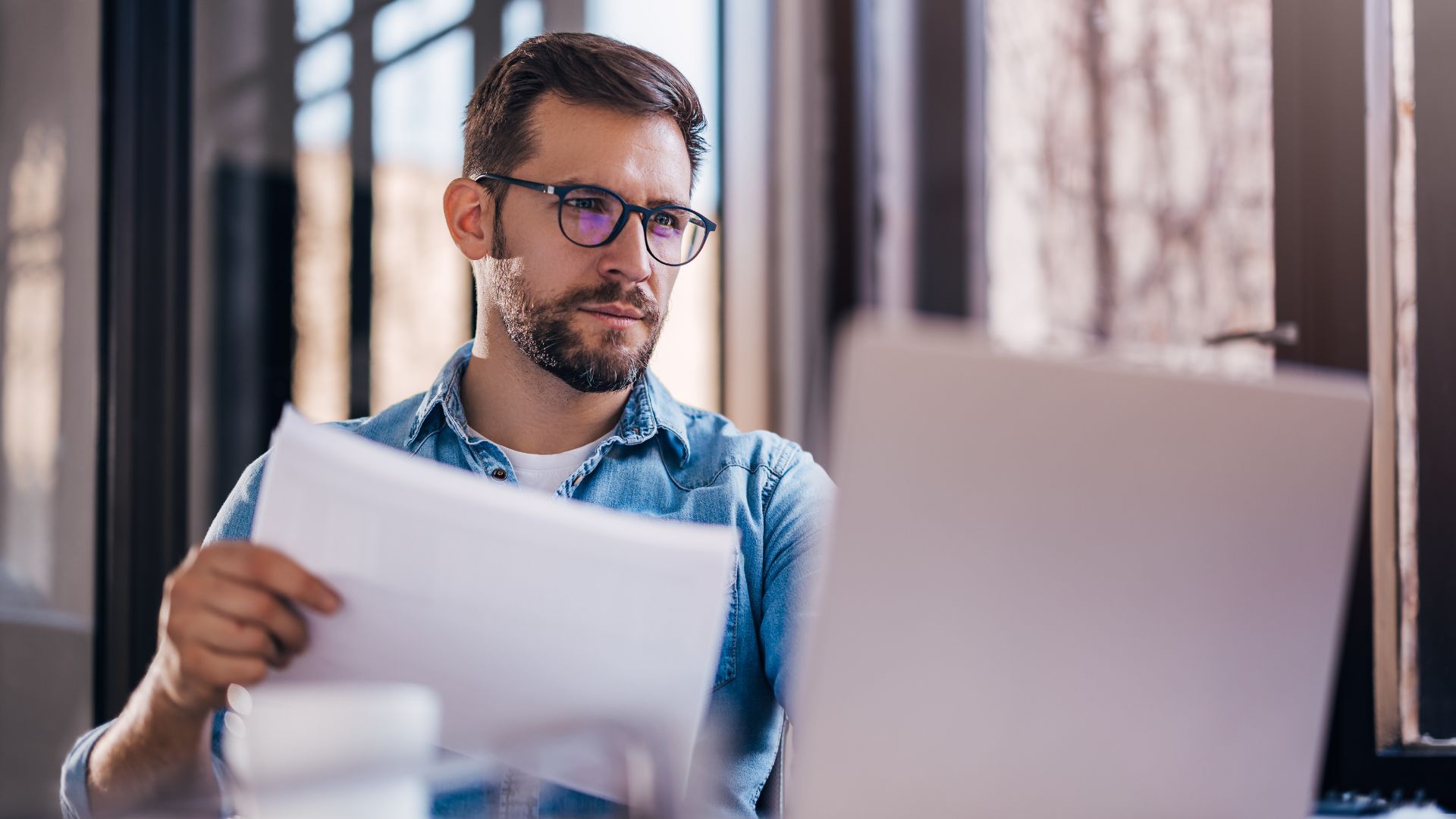 A man wearing glasses sits at a desk, reviewing paperwork while looking at a laptop screen, possibly researching legal or financial matters such as handling an insolvent estate.