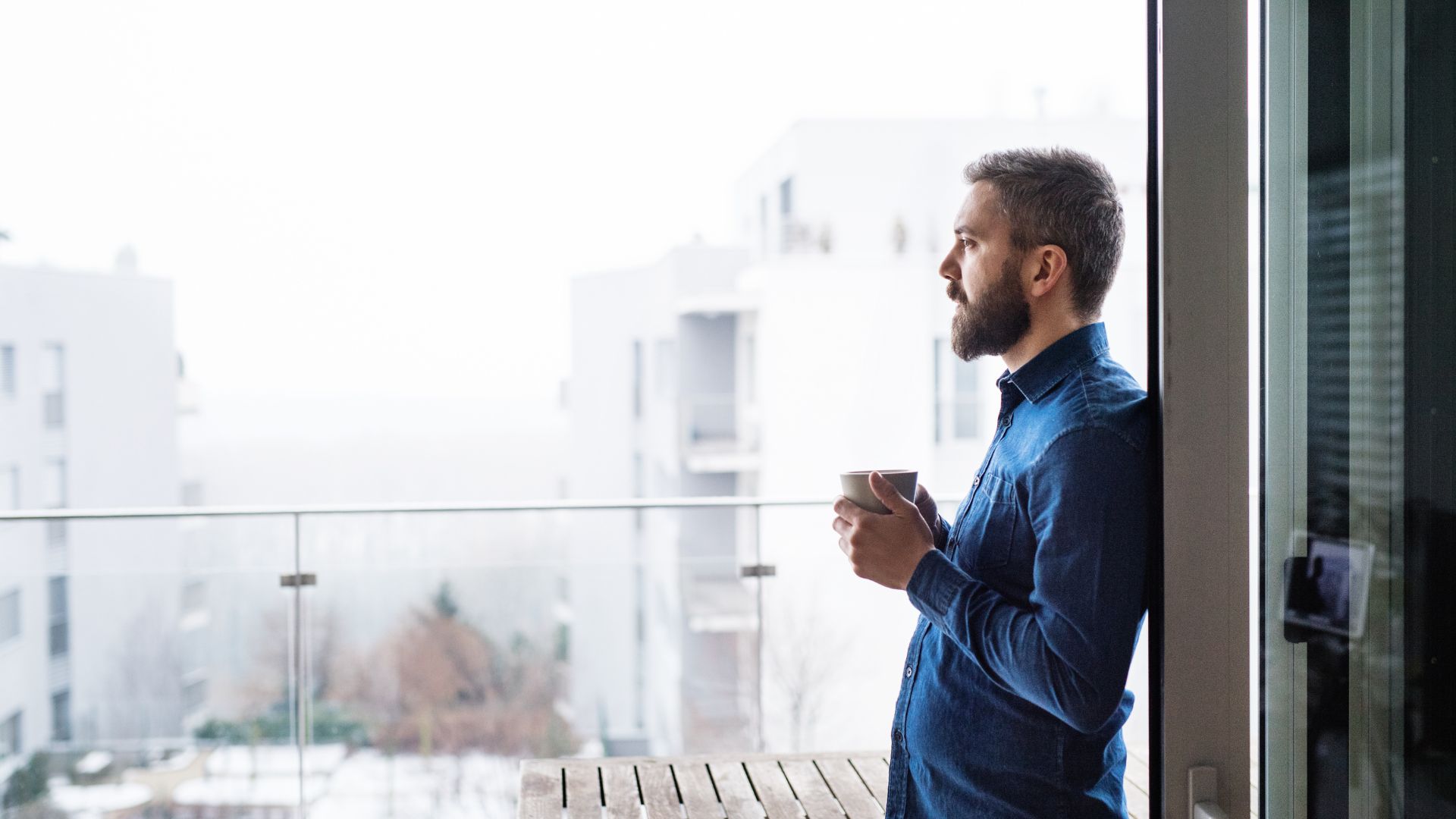 A man stands on a balcony holding a cup, gazing thoughtfully at the view of modern apartment buildings on a cloudy day—possibly reflecting on major life changes such as going through a divorce in Nevada or making important legal decisions.